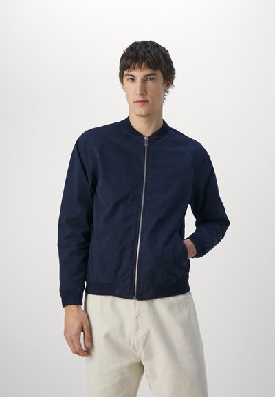 Chaqueta bomber azul marino con cremallera frontal, cuello y puños acanalados. Textura suave, cuenta con dos bolsillos laterales. El modelo lleva pantalones de color claro.