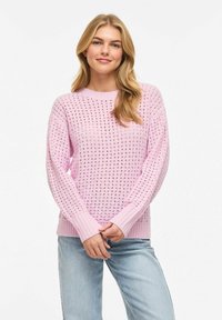 Jeune femme aux longs cheveux blonds portant un pull rose tricoté avec des trous et un jean bleu clair, debout devant un fond blanc.