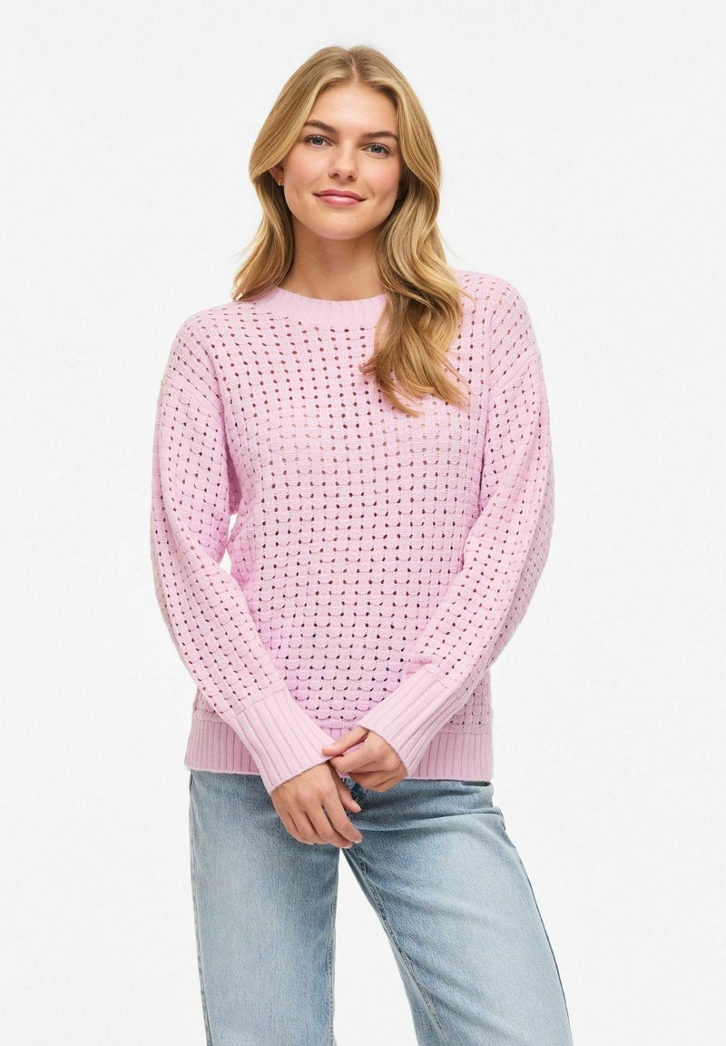 Jeune femme aux longs cheveux blonds portant un pull rose tricoté avec des trous et un jean bleu clair, debout devant un fond blanc.