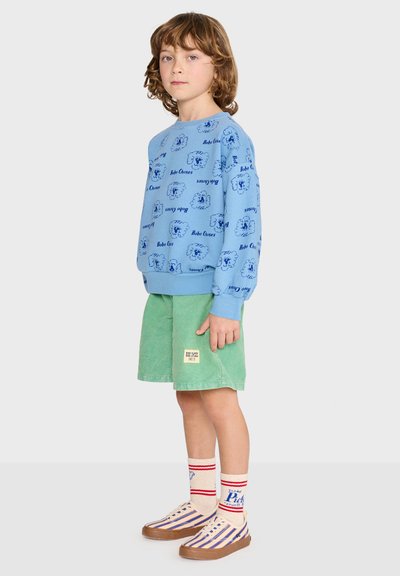 Enfant aux cheveux ondulés portant un sweat-shirt bleu à motifs, un short vert, des chaussettes rayées et des baskets multicolores, debout de profil.