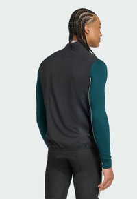 Black vest with a high collar, mesh paneling, and elastic hem, worn over a teal long-sleeve shirt and black fitted pants.