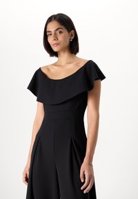 Melna kombinezona bez pleciem ar izteiktu dekoltē, strukturētu augšējo daļu un platām, krokām rotātām biksēm. Izvēlēts gluds audums un pielāgota piegriezuma forma.