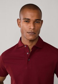 Bordová polo košeľa s klasickým golierom, tromi čiernymi gombíkmi a hladkou textúrou. Jednoduchý dizajn, priliehavý strih a bez vzorov.