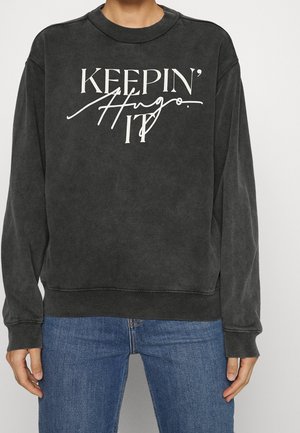 Felpa nera a maniche lunghe con collo tondo con scritta bianca "KEEPIN' IT" e scritta corsiva "Hugo", indossata con jeans blu.