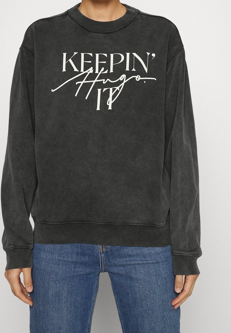 Sweat noir à manches longues avec col rond, portant le texte blanc « KEEPIN' IT » et le texte en cursive « Hugo », porté avec un jean bleu.