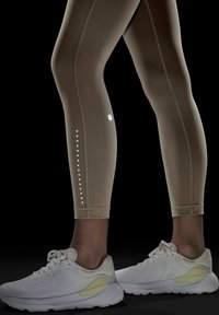 Beige träningsleggings med reflekterande prickiga detaljer, med hög midja och kontrasterande sömmar, matchade med vita sneakers.