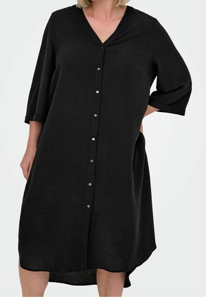 Kvinde iført løs sort knap-op kjole med trekvartærmer og V-hals, stående med den ene hånd i lommen.