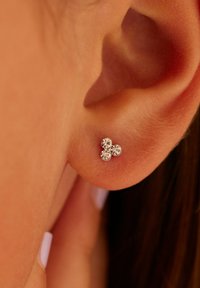 Zilveren stud oorbellen met drie ronde rhinestones, gerangschikt in een driehoekig patroon tegen een close-up van een oor. Gladde textuur.