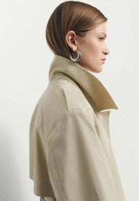 Trench coat bej croppat cu guler înalt, realizat dintr-un material neted. Cercei din argint în formă de cerc adaugă un accent metalic. Vedere din profil.