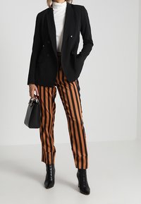 Czarny podwójnie zapinany żakiet na białym golfie, zestawiony z brązowo-czarnymi spodniami w paski i czarnymi botkami za kostkę. Czarna torebka w ręku.