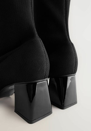 Mustad musta lõngaga pahklutsa saapad, millel on plätseeritud musta läikiva kandiga ruudukujuline vorm. Tekstuur on sile ja kogu disain on stiilne ja kaasaegne.