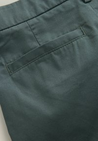 Gros plan d’un pantalon en tissu vert montrant une poche arrière horizontale avec des coutures apparentes et une couture à la taille.