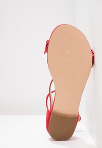Punaiset tasapohjaiset sandaalit nilkkaremmillä, joissa näkyy sileä beige pohja ja teksturoitu ruskea korko valkoista taustaa vasten.