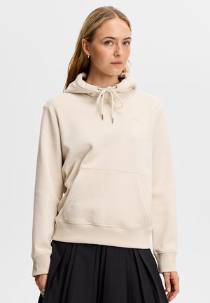 Kvinde med langt blondt hår iført en lys beige hættetrøje med frontlomme og sort plisseret nederdel, stående mod en ensartet hvid baggrund.