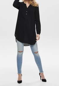 Czarna tunika z długim rękawem i dekoltem w kształcie V, zdobiona guzikami, w połączeniu z jasnoniebieskimi, postrzępionymi rurkami i czarnymi butami na wysokim obcasie.