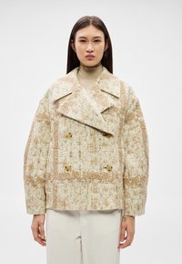 Sieviete valkā bēša un krēmkrāsas teksturētu dubultpogājamu žaketi ar lieliem atlokiem virs bēša augstu uzrullēta kakla džempera un gaišām biksēm.