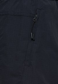 Svart tyg med ett rutmönster, utrustat med en dragkedjeficka och justerbar snörning. Slät yta med förstärkta sömmar.