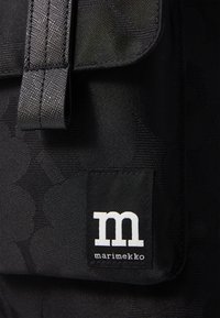Mustaa kangaskassia, jossa on teksturoitu kuvio, läppäsuljin ja näkyvä valkoinen etiketti, jossa lukee "marimekko" ja tyylitelty "m"-logo.