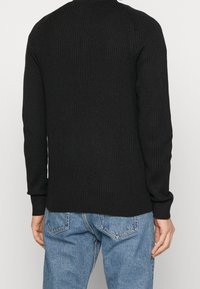 Svart ribbad stickad tröja med långa ärmar och rund hals, utrustad med ett texturerat tyg och åtsittande mudd, bärs över ljusblå jeans.