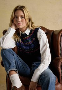 Jūras spēku zils adīts veste ar daudzkrāsainiem ģeometriskiem rakstiem, uzvilkta virs baltas krekla. Modelis sēž uz brūna ādas krēsla, uzvelkot džinsus.