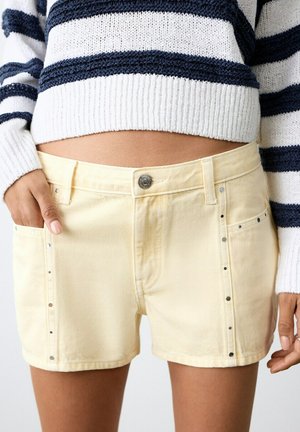 Personne portant un short en denim jaune pâle avec des rivets argentés et un pull à rayures bleu marine et blanc, la main dans la poche.