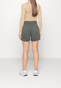 Pantalones cortos de color verde oscuro con un ajuste holgado, dos bolsillos traseros y una cinturilla. Combinados con una camiseta de manga larga color beige y zapatillas de color claro.