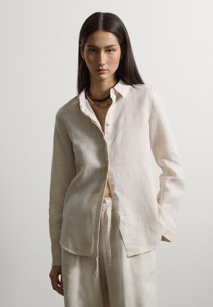 Frau mit glattem, langem, dunklem Haar, die ein aufgeknöpftes cremefarbenes Leinenhemd, passende Hosen und geschichtete Perlenketten trägt, vor einem schlichten Hintergrund steht.