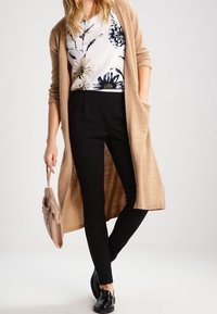 Cardigan lungo beige, top a fiori bianco e blu, pantaloni neri aderenti e scarpe nere. Tiene in mano una borsa leggera rosa.