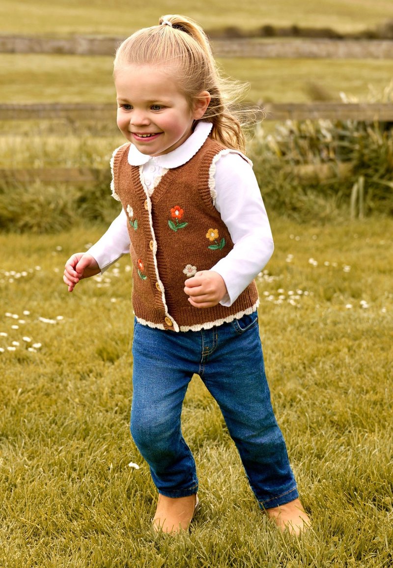 Chaleco de punto marrón con bordado floral, camisa blanca con cuello, jeans azules y pies descalzos, con hierba de fondo. Niño en movimiento.