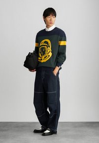 Σκούφος σε σκούρο πράσινο χρώμα με κίτρινο γραφικό αστροναύτη και ρίγες στα μανίκια, σε συνδυασμό με φαρδιά τζιν παντελόνια και μαύρα αξεσουάρ.