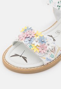 Białe sandały z kwiatowym wzorem, ozdobione różowymi, żółtymi i niebieskimi skórzanymi kwiatami oraz srebrnymi ćwiekami; brązowa podeszwa z przeszyciem.