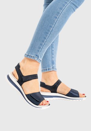 Feet wearing navy blue velcro sandals paired with light blue skinny jeans, toes painted red, against a light gray background.