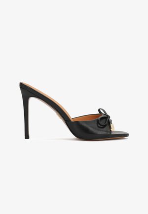 Mules de tacón alto de cuero negro con punta abierta, con un lazo decorativo y detalles de herrajes dorados en el lateral. Textura suave y diseño elegante.