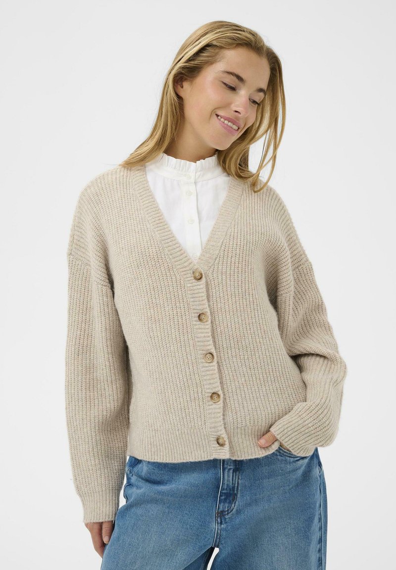 Beige strikket cardigan med V-hals, knaplukning og ribbet tekstur. Har lange ærmer og en afslappet pasform, parret med lyseblå jeans.