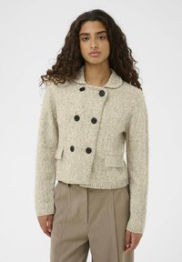 Pletena bež jakna s teksturiranim završetkom, dvostruko zakopčavanje, veliki crni gumbi i ovratnik. Ima dva prednja džepa.