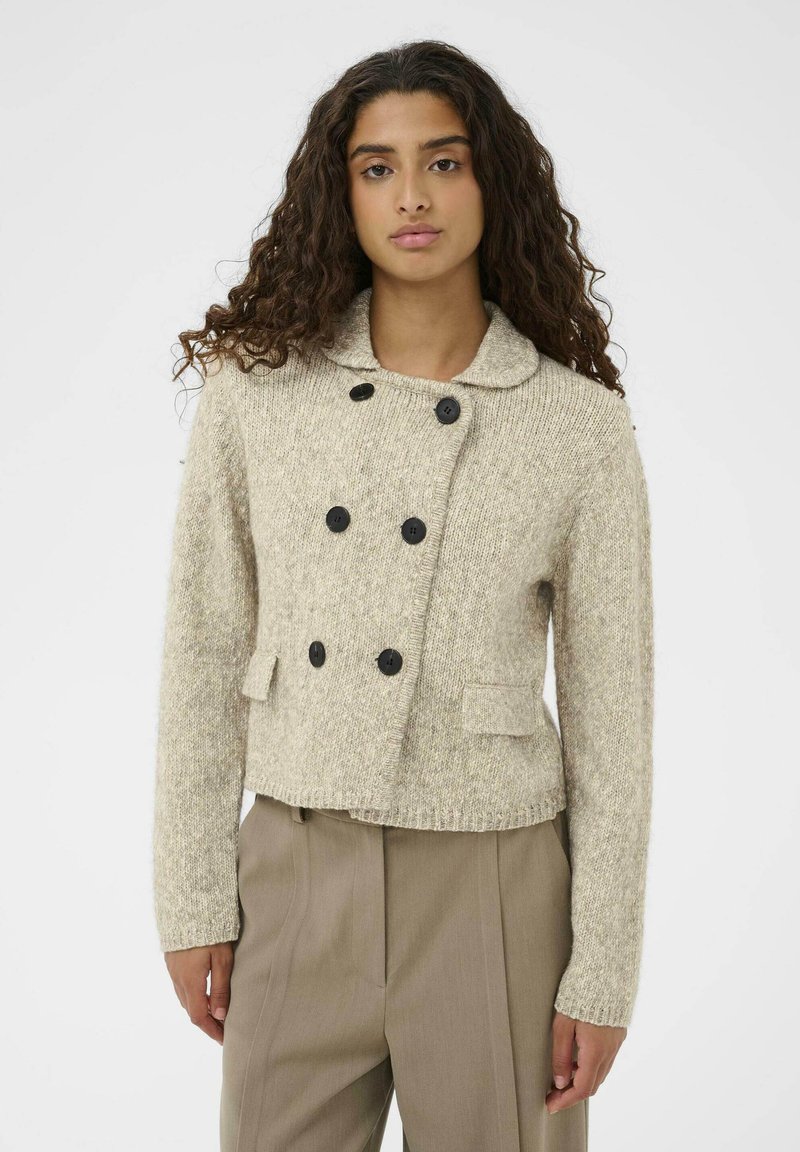 Pletena bež jakna s teksturiranim završetkom, dvostruko zakopčavanje, veliki crni gumbi i ovratnik. Ima dva prednja džepa.