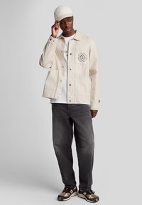 Lys beige jakke med to forlommer, sort logo-detalje, parret med en hvid T-shirt, grå jeans og mønstrede sko.