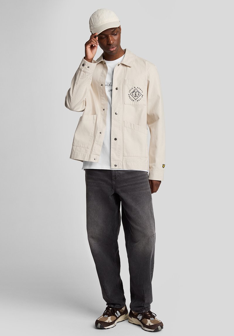 Lys beige jakke med to forlommer, sort logo-detalje, parret med en hvid T-shirt, grå jeans og mønstrede sko.