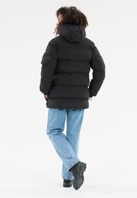 Person der går væk iført en sort hættejakke med fyld, lys blå jeans og sorte støvler på en hvid baggrund.