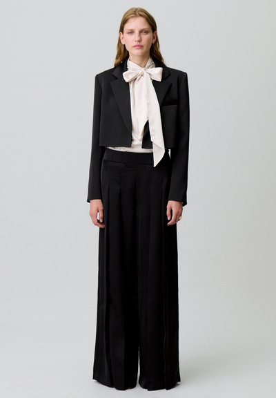Blazer noir court avec une poche, porté sur une blouse blanche avec un grand nœud. Pantalon noir taille haute et plissé.