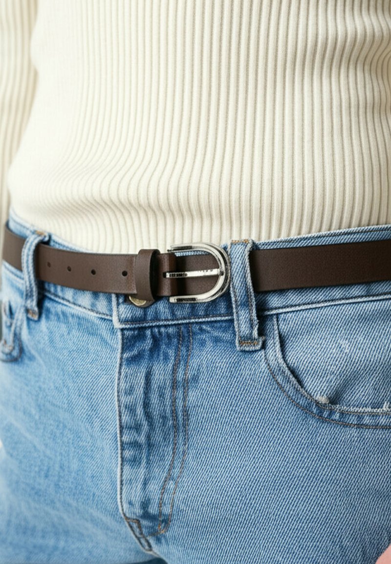 Bruine leren riem met een zilveren rechthoekige gesp, gedragen over lichtblauwe denim jeans. Geribbeld wit shirt zichtbaar boven de riem.