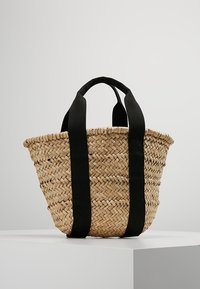 Borsa a tracolla in paglia intrecciata con manici larghi in tessuto nero, in piedi su un piedistallo bianco contro uno sfondo semplice.