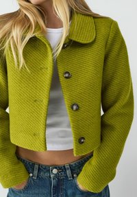 Veste cropped texturée verte avec un design côtelé, dotée d'un col et de boutons noirs, associée à un haut blanc et un jean en denim.