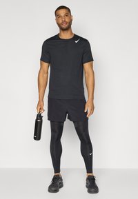 T-shirt de sport noire avec un design texturé, shorts noirs, leggings noirs et baskets noires, tenant une bouteille d'eau noire.