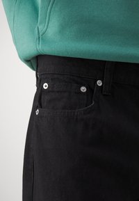 Black denim pants with silver hardware, featuring a front pocket and a smooth texture, paired with a green sweatshirt.