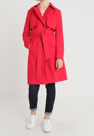 Personne portant un trench-coat rouge jusqu'aux genoux avec une ceinture, un jean foncé retroussé et des baskets blanches sur un fond uni.