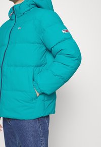 Tealfärgad pufferjacka med dragkedja fram, huva och elastiska manschetter. Har en logotyp på ärmen och sidofickor. Slät, glänsande textur.