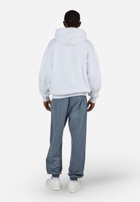 Λευκή υπερμεγέθη κουκούλα με κουκούλα, σε συνδυασμό με φαρδιά γκρίζα φόρμα. Το σύνολο διαθέτει ραμμένες μανσέτες και λευκά αθλητικά παπούτσια.