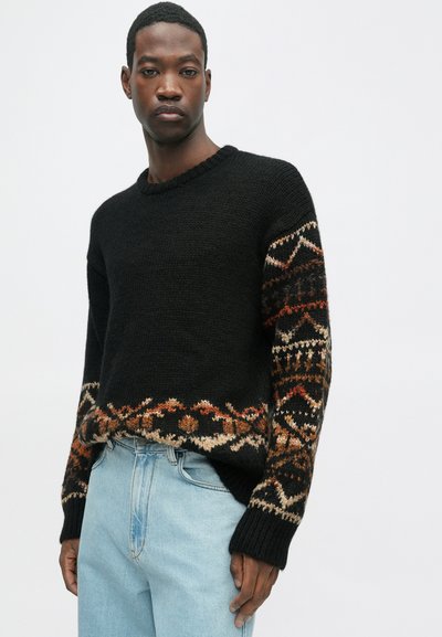 Fekete kötött pulcsi mintás ujjakkal, meleg színekben. Bordás mandzsetták és szegély. Világoskék farmernadrággal párosítva.