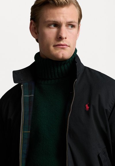 Črna zip-up jakna s karirastim podložjem in rdečim logotipom, oblečena čez temno zeleno turtleneck pulover s teksturirano pletenino.